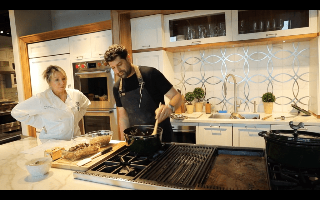 Two people stand in a modern kitchen showroom; one stirs food in a pot on the stove while the other looks on, with various ingredients and utensils displayed on the counter.
