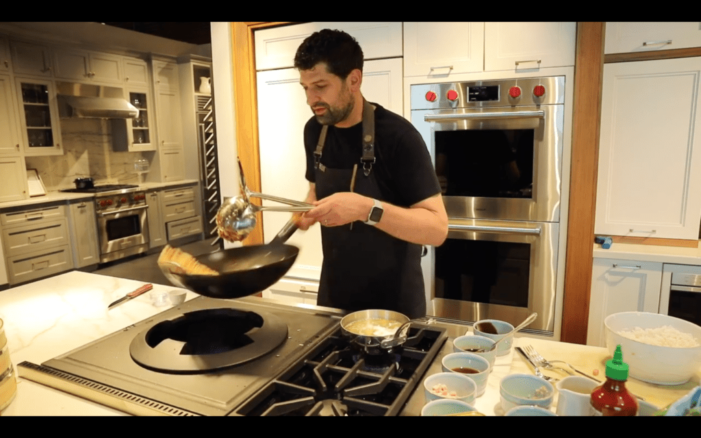 A man in a kitchen holds a wok and metal tongs over a wolf appliance stove, preparing food with several small bowls of ingredients arranged on the counter.