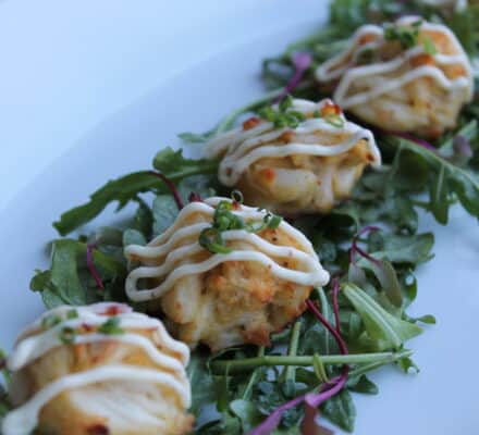 Four small crab cakes topped with a drizzle of sauce are arranged on a bed of mixed greens on a white plate—a dish worthy of any luxury kitchen appliance showroom.
