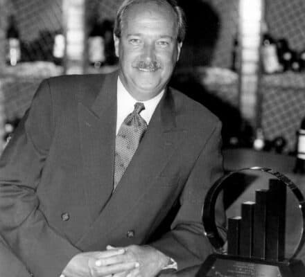 A man in a suit stands beside an award trophy on a counter, with wine bottles on shelves in the background of a luxury kitchen appliance showroom.