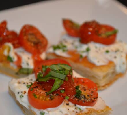 Three pieces of puff pastry topped with sliced cherry tomatoes, creamy cheese, fresh basil, and black pepper are artfully arranged on a white plate—perfect inspiration for your kitchen showroom or those seeking a luxury kitchen appliance experience.