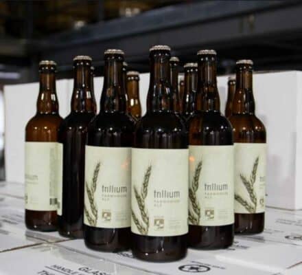 Several glass beer bottles with “Trillium Farmhouse Ale” labels are arranged on stacked white cardboard boxes in an indoor setting, the perfect touch for any gathering in a luxury kitchen featuring Wolf or Sub-Zero appliances.