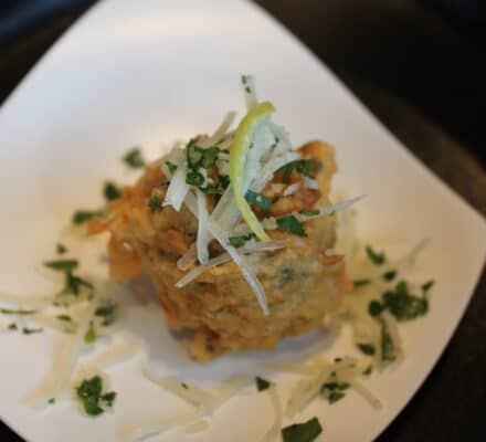 A plated fried appetizer garnished with grated cheese, chopped herbs, and thin lemon slices is artfully presented on a white dish—perfect for serving in a luxury kitchen alongside your favorite Sub Zero appliance.