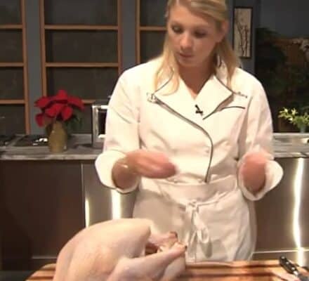 A chef in a white coat stands behind a kitchen counter, preparing to work with a raw turkey placed on a wooden cutting board, with a luxury kitchen appliance from Wolf Appliance showcased in the background.