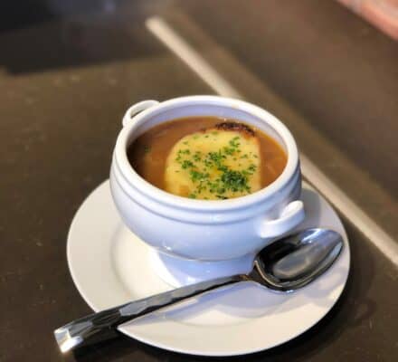 A white bowl of French onion soup with a slice of toasted bread and melted cheese on top, served on a white plate with a spoon—perfectly crafted in a kitchen showroom featuring the latest Sub Zero appliances.