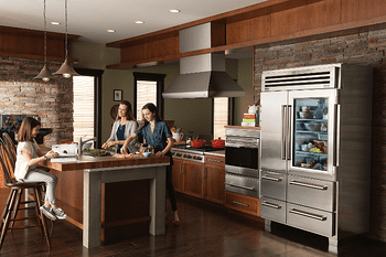 Three people are in a modern kitchen; two are preparing food at the counter while one sits on a stool, with luxury kitchen appliances and stainless steel finishes visible in the background.
