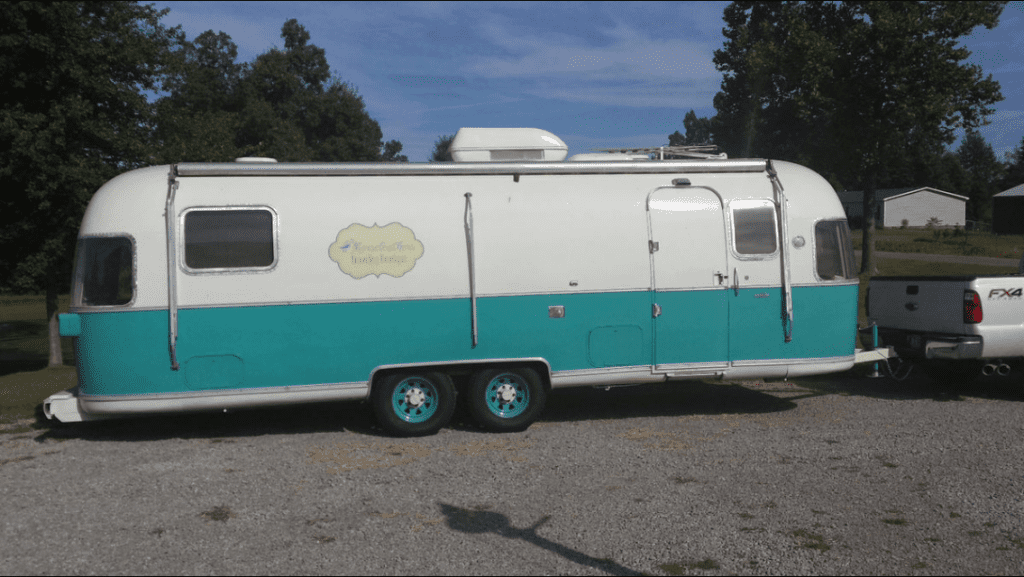 A vintage turquoise and white travel trailer hitched to a white pickup truck, parked on gravel with grass and trees in the background—its cozy interior reminiscent of a kitchen showroom featuring stylish Wolf appliances.