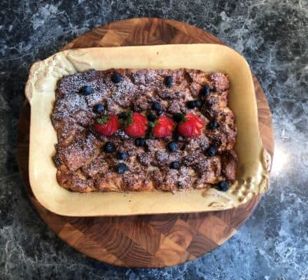 A rectangular baked dessert topped with powdered sugar, blueberries, and three strawberries, served in a ceramic dish on a wooden board—perfect for showcasing in a kitchen showroom featuring luxury kitchen appliances.
