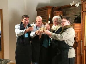 Five adults wearing aprons stand in a luxury kitchen, clinking glasses in a toast, smiling and celebrating together beside a sleek Sub Zero appliance.