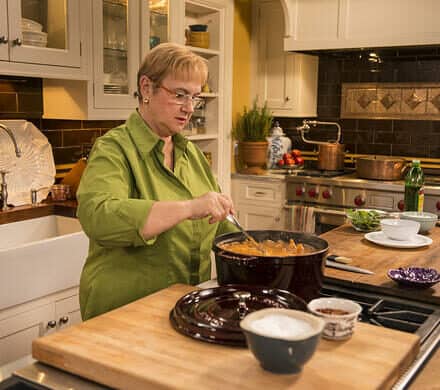 A woman in a green shirt stirs food in a pot on a stove next to a sleek Wolf appliance in a well-lit, modern kitchen with wooden countertops and white cabinets.