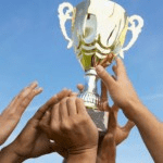 Multiple hands lifting a gold trophy cup against a clear blue sky, symbolizing teamwork and achievement—much like the collaboration found in a luxury kitchen appliance showroom.