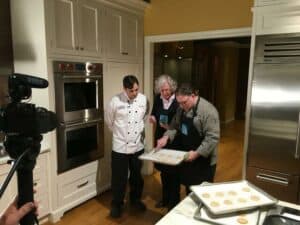 Three people in a kitchen showroom examine trays of unbaked cookies; one wears a chef’s coat, the other two aprons. A camera is set up in the foreground, capturing the baking session among premium appliances.