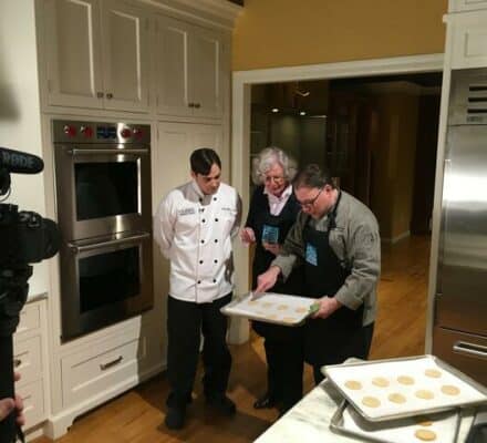 Three people in a kitchen showroom examine trays of unbaked cookies; one wears a chef’s coat, the other two aprons. A camera is set up in the foreground, capturing the baking session among premium appliances.