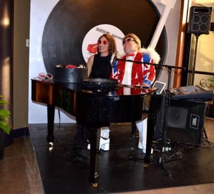 Two people in costumes stand behind a black piano at an appliance showroom; one wears red sunglasses and a feathered jacket, with a large vinyl record prop in the background.