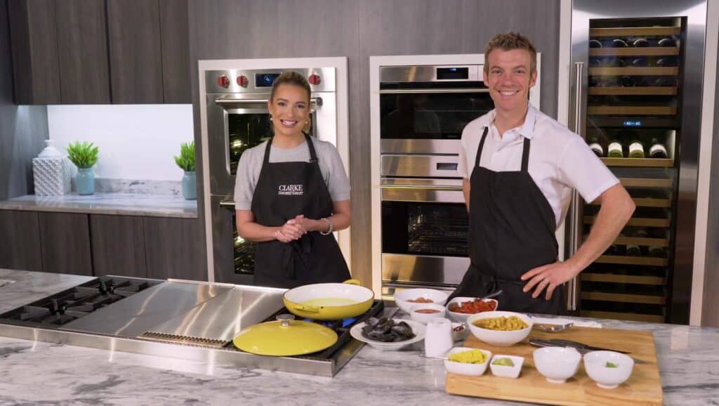 Two people wearing aprons stand in a modern kitchen, smiling at the camera, with cooking ingredients and utensils arranged on the counter in front of a gleaming Sub-Zero appliance.