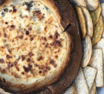 A round bread bowl filled with baked cheesy spinach artichoke dip, surrounded by assorted crackers arranged in a circular pattern.