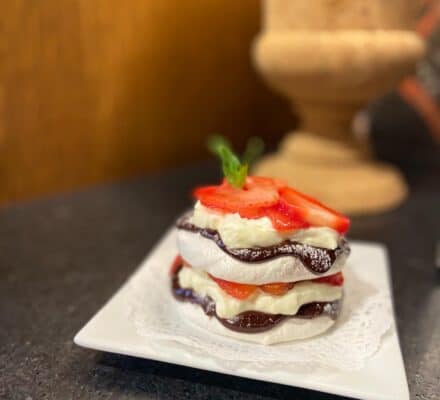 A dessert with layers of meringue, whipped cream, chocolate sauce, and sliced strawberries, garnished with a mint leaf, served on a square white plate—perfect for impressing guests in your modern kitchen showroom.