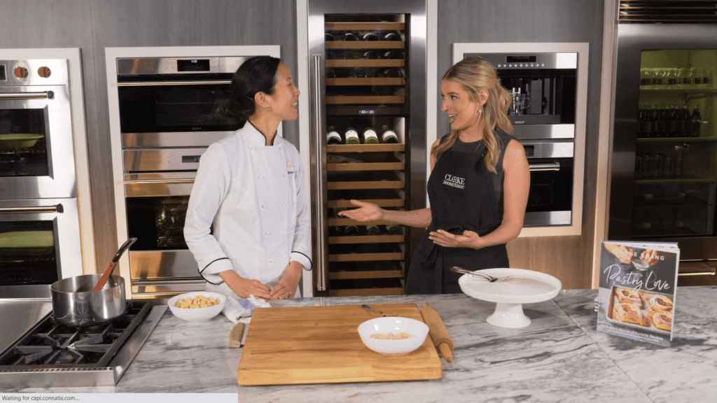 Two women stand in a modern kitchen showroom, one in a chef’s jacket and the other in a black apron, chatting beside a counter with kitchenware, ingredients, and a sleek Wolf appliance.