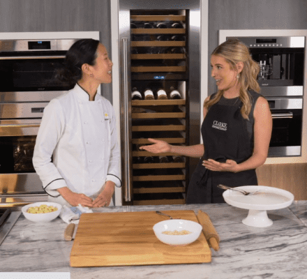 Two women stand in a modern kitchen showroom, one in a chef’s jacket and the other in a black apron, chatting beside a counter with kitchenware, ingredients, and a sleek Wolf appliance.