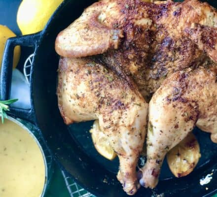 Roast spatchcock chicken seasoned with herbs in a cast iron skillet, surrounded by lemon halves, prepared to perfection on a Wolf appliance, with a pot of sauce and fresh rosemary nearby.