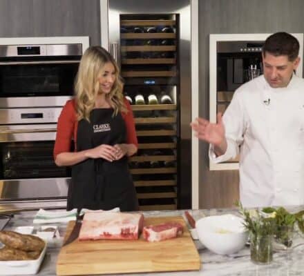 A woman in an apron and a chef in a white coat stand behind a kitchen counter with raw meat, potatoes, and herbs, surrounded by sleek sub zero appliances in a modern kitchen showroom.