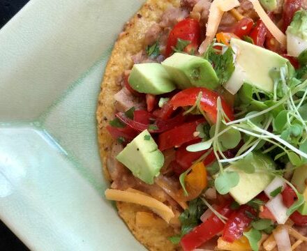 A close-up of a tostada topped with avocado, tomatoes, shredded cheese, microgreens, and other fresh vegetables on a green plate in front of a luxury kitchen appliance.