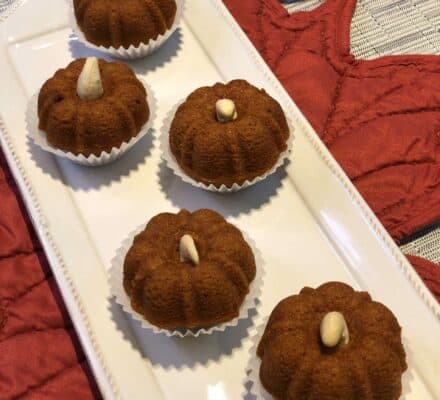 Six round, brown mini cakes with a single nut on top, arranged neatly on a rectangular white plate in what could be an elegant kitchen showroom with red mats in the background.