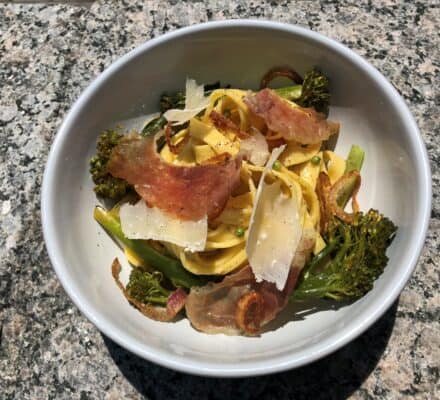 A bowl of pasta topped with shaved parmesan, crispy prosciutto, broccolini, and fried shallots sits on a granite surface—perfect inspiration for any appliance showroom or luxury kitchen appliance display.