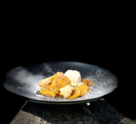A black plate with a serving of peach cobbler and a scoop of vanilla ice cream sits on a stone surface, evoking the sophistication of a luxury kitchen appliance display against a striking black background.