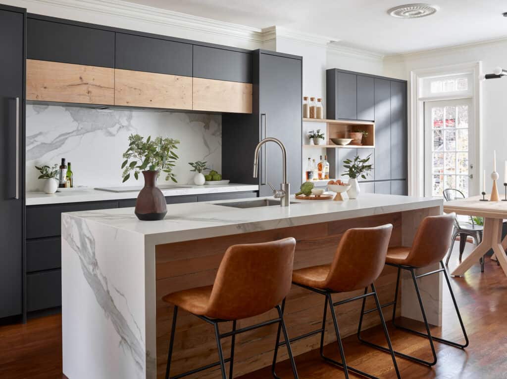 Modern kitchen with a marble island, brown leather bar stools, matte black cabinets, and wood accents. Featuring premium Wolf appliances, plants and kitchenware are placed on the counters and open shelves.