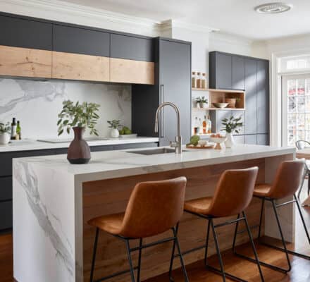 Modern kitchen with a marble island, brown leather bar stools, matte black cabinets, and wood accents. Featuring premium Wolf appliances, plants and kitchenware are placed on the counters and open shelves.