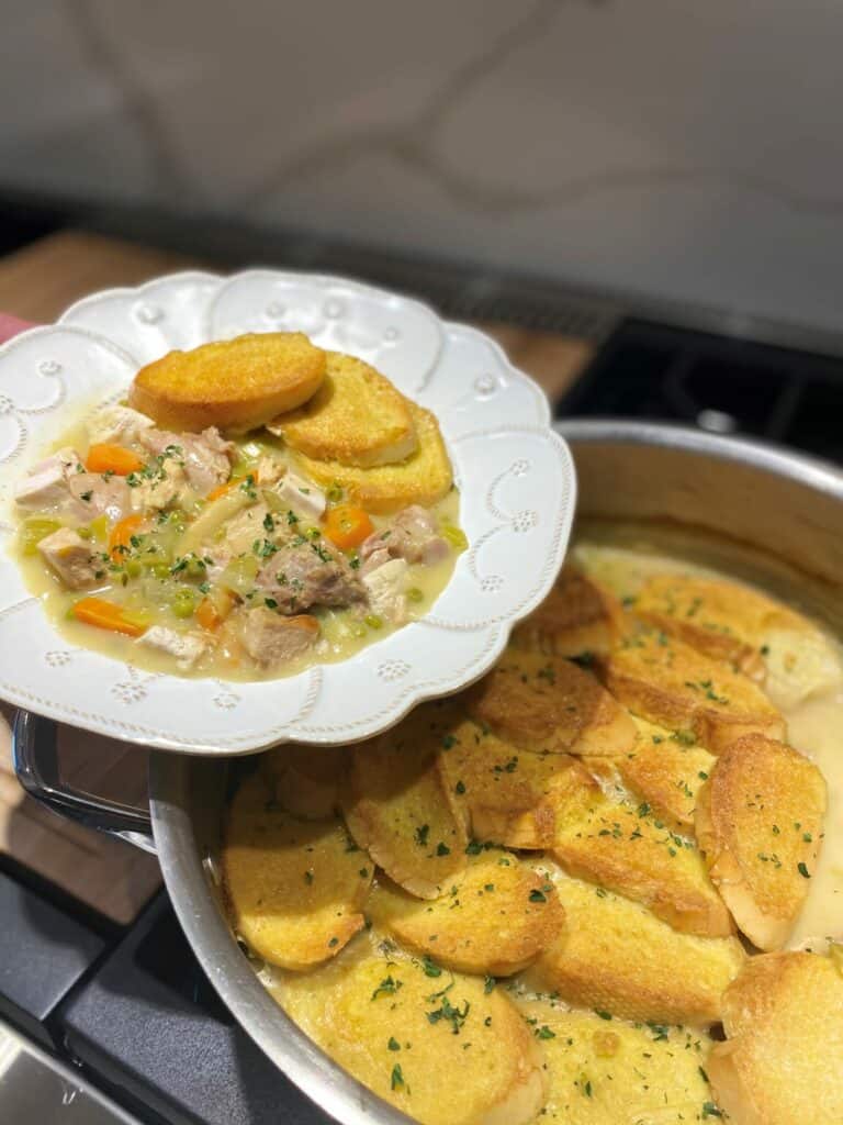 A white plate holds a serving of chicken stew with vegetables and toasted bread rounds, next to a pan filled with more stew topped with toasted bread slices—an inviting scene perfect for any kitchen showroom display.