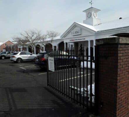 A black iron gate opens to a parking lot with several cars in front of a white building labeled "CLARKE," renowned as a kitchen showroom for luxury kitchen appliance brands, on a snowy day.