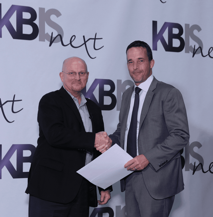 Two men in business attire shake hands at a KBIS Next event, standing in front of a branded backdrop, possibly discussing the latest Wolf appliance for a luxury kitchen showroom, with one holding a sheet of paper.