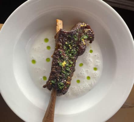 A plate with a bone-in piece of cooked meat garnished with herbs, served on foamy white sauce with green oil drops—just the kind of dish you'd find showcased in a luxury appliance showroom featuring Sub Zero and Wolf appliance displays.