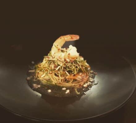 A plated dish of pasta topped with a single shrimp and herbs is elegantly presented on a black plate against a dark background, evoking the refined style of a luxury kitchen showroom with Wolf and Sub-Zero appliances.