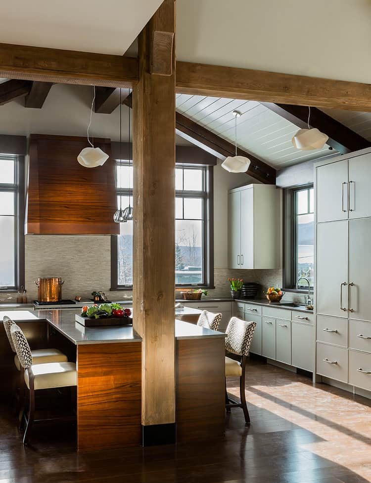 Modern kitchen showroom with high ceilings, exposed wood beams, pendant lights, an island with seating, and a mix of wood and white cabinetry. Sunlight streams through large windows, highlighting premium Wolf appliances.