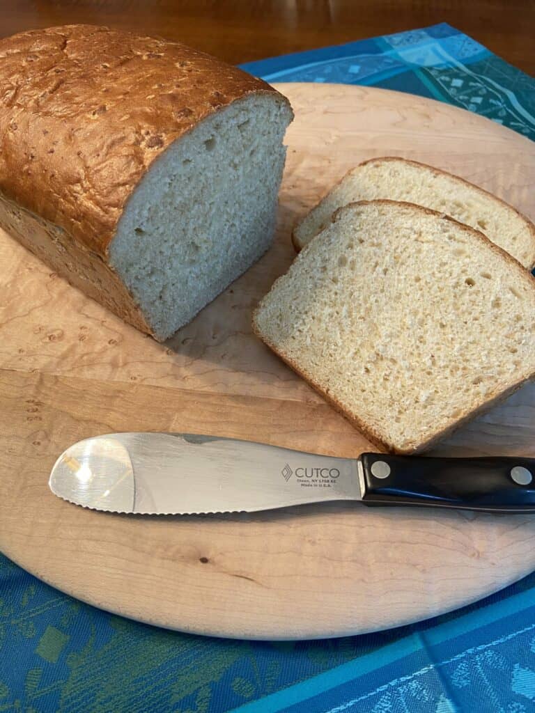 A loaf of bread with two slices cut, placed on a wooden cutting board next to a serrated Cutco knife, creates the perfect scene in front of a gleaming Wolf appliance—ideal for any luxury kitchen appliance lover.