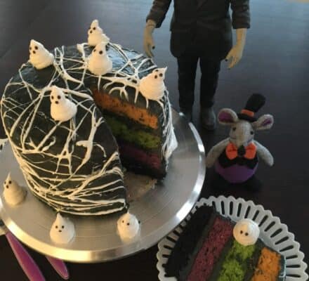 A Halloween-themed cake with ghost decorations and colorful layers sits on a table in a kitchen showroom, joined by a Frankenstein figure and a mouse doll dressed in purple.