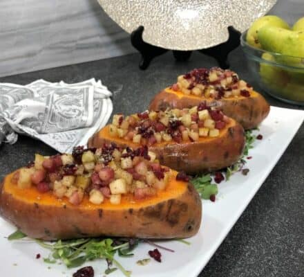Three halved baked sweet potatoes topped with diced apples, cranberries, and nuts are arranged on a white rectangular plate, styled as they might appear in an elegant appliance showroom display. Pears and a napkin are visible in the background.