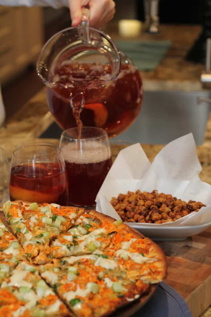 A person pours a red beverage from a glass pitcher into a glass beside a pizza, two filled glasses, and a bowl of roasted chickpeas on a kitchen counter with a luxury kitchen appliance in view.