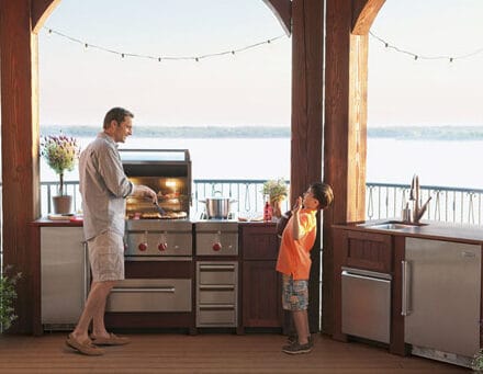 An adult grills food on a luxury kitchen appliance in an outdoor kitchen while a child stands nearby, with a lake and trees visible in the background.