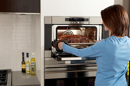 A person places a baking tray with a cooked roast into a built-in Wolf appliance oven in a modern kitchen. Bottles of oil and vinegar are on the countertop nearby.