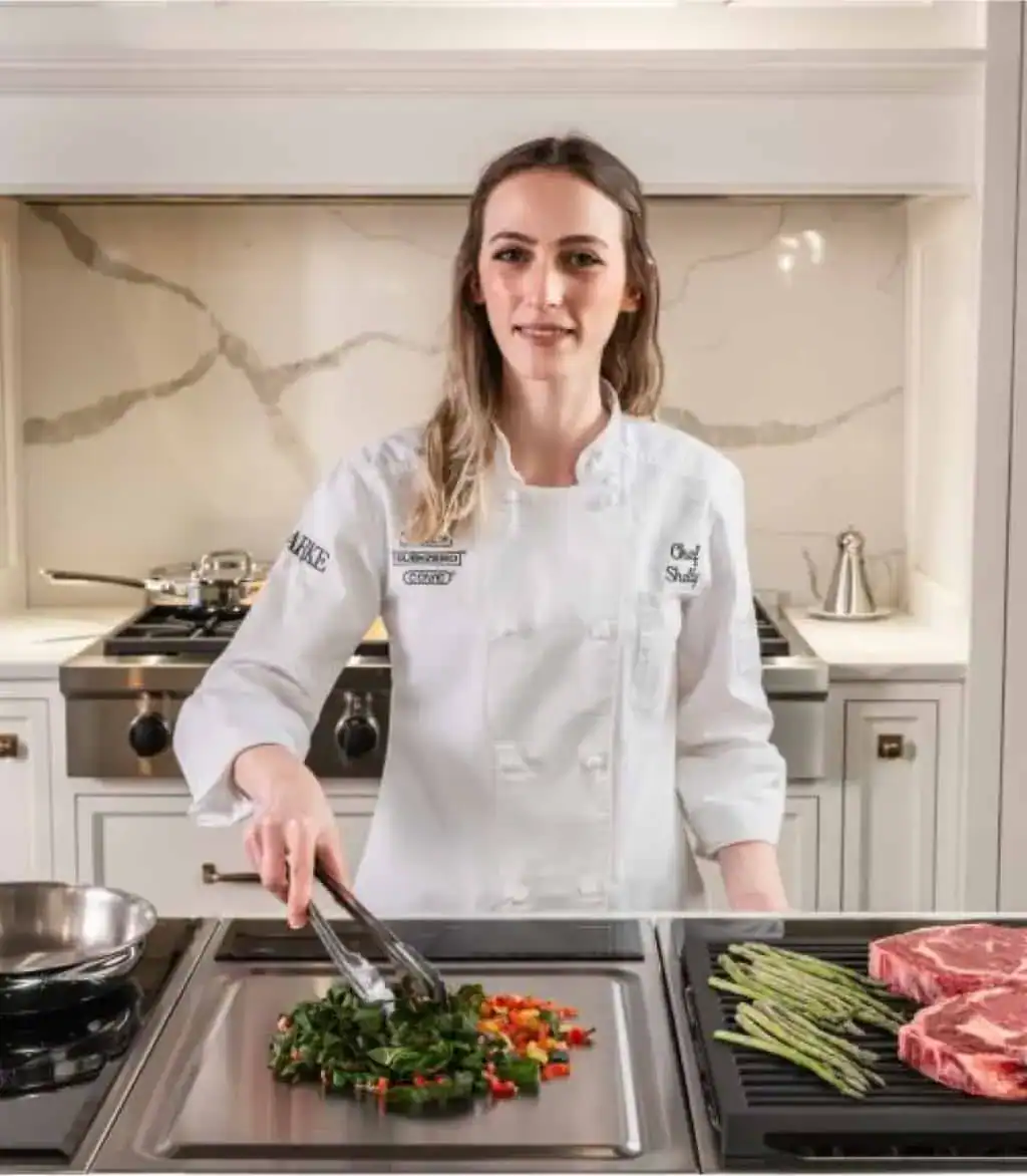 A chef in a white uniform cooks vegetables on a Wolf appliance stovetop with tongs; asparagus and steaks are prepared nearby in a modern kitchen featuring premium Sub-Zero appliances.