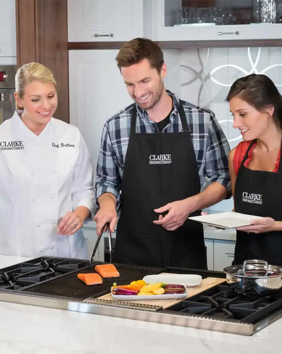 Three people, two women and one man, stand around a stove in an appliance showroom as the man grills salmon and vegetables on a Wolf appliance. All wear aprons; one woman wears a chef coat labeled "Clarke.