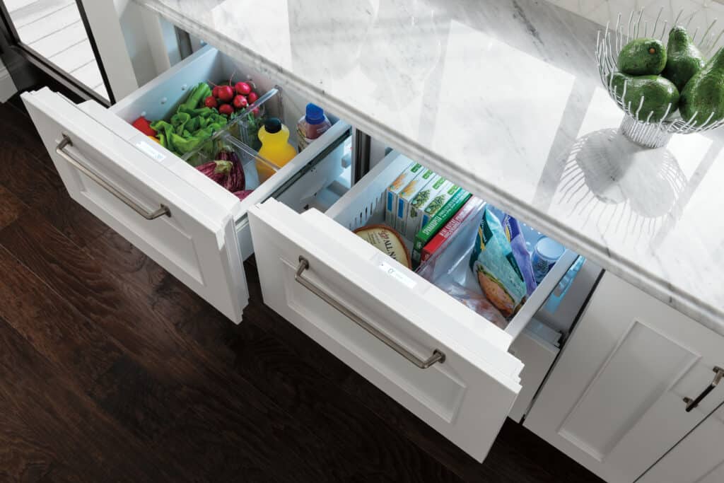 Two open refrigerator drawers beneath a marble countertop display organized food items, perfect for a luxury kitchen appliance in a modern kitchen showroom. Avocados sit in a bowl on the counter above.