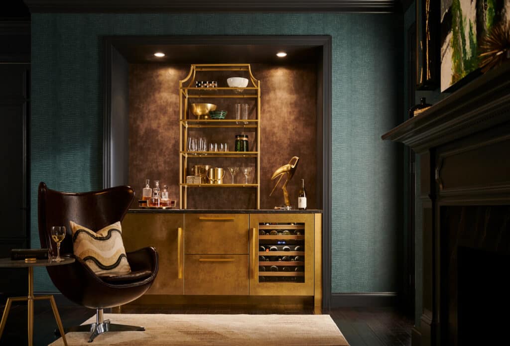 A modern home bar with gold cabinets, open shelves displaying glassware, a wine fridge, luxury kitchen appliances, bottles, and a sculptural lamp beside a leather chair and side table in a dimly lit room.