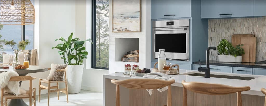 Modern kitchen and dining area with light wood and blue cabinetry, built-in oven, island seating, large windows, indoor plants, and neutral decor—perfect inspiration for your next visit to a kitchen showroom.