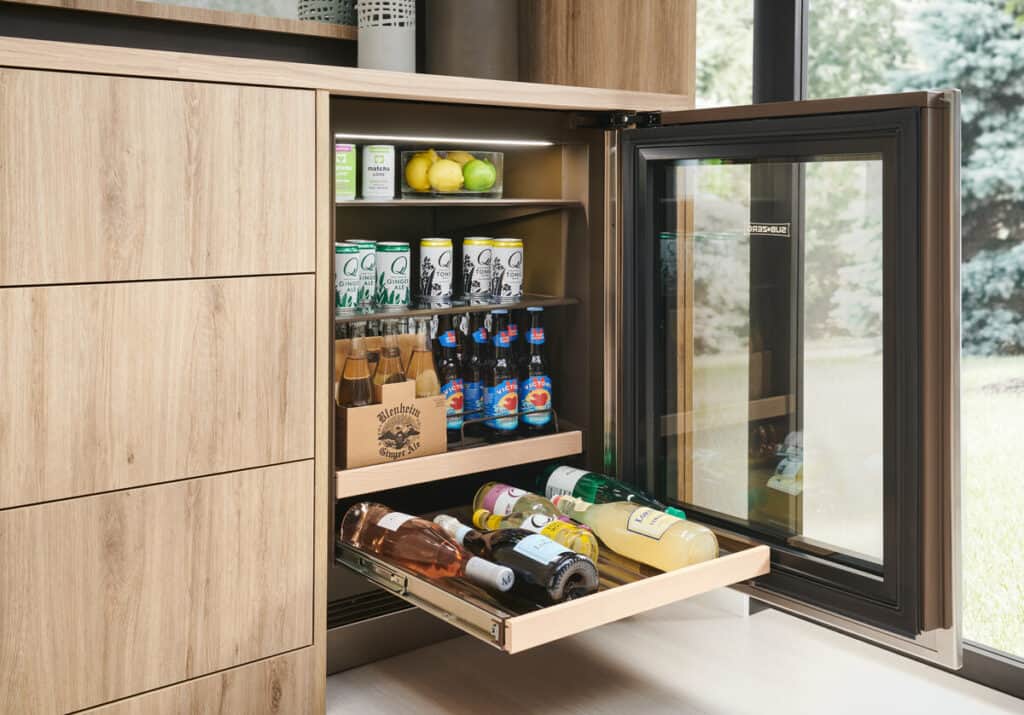 A small built-in fridge with the door open, showing shelves stocked with cans, bottles, lemons, and limes—an ideal addition to any kitchen showroom displaying the latest in luxury kitchen appliance design beside a window with a view of greenery.