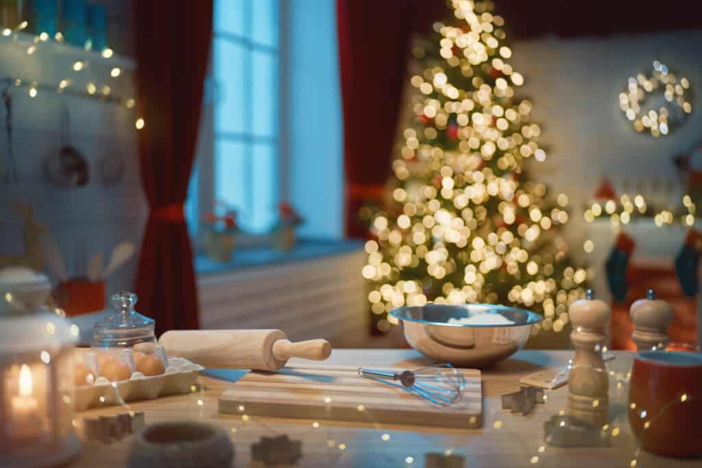 A kitchen counter with baking tools and ingredients in the foreground, featuring a luxury kitchen appliance, with a decorated Christmas tree and festive lights blurred in the background.